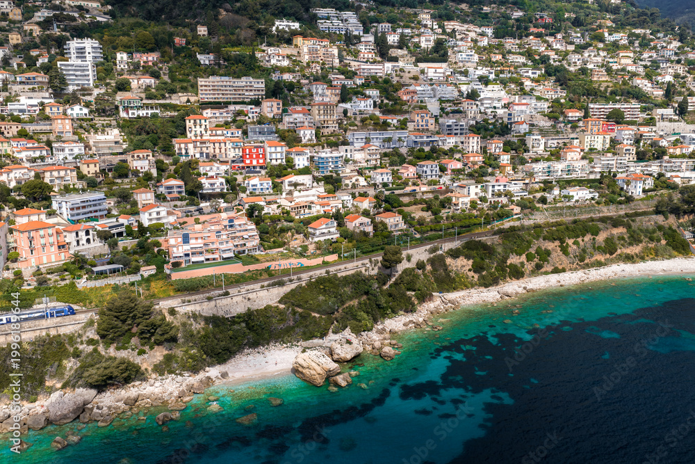 Fototapeta premium Aerial view of a terraced town near Monaco on the French Riviera with hillside villas, a seaside road and rail line with a train, rocky coves, and turquoise water at midday.