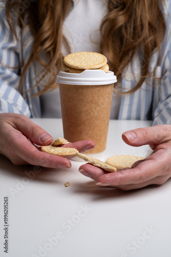 Woman holding broken cookie near paper coffee cup on white table. Snack break concept. 