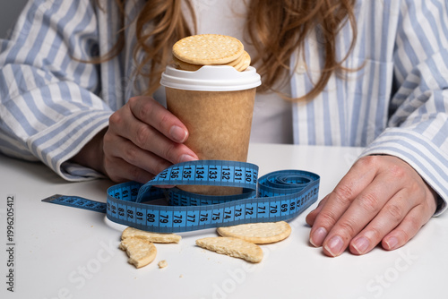 Woman holding coffee cup with cookies and blue measuring tape on table. Dietary control concept. 