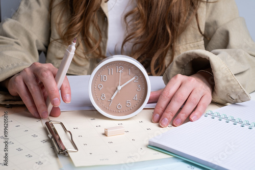 Woman holding alarm clock and writing in notebook with calendar on desk. Effective time management concept. 