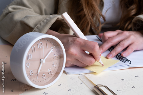 Woman holding alarm clock and writing in notebook with calendar on desk. Effective time management concept. 