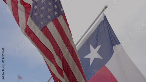 A cinematic shot of the United States national flag and the Texas state flag (Lone Star Flag) flying side-by-side on flagpoles. Both flags billow in a steady breeze against a clear, vibrant blue sky.
