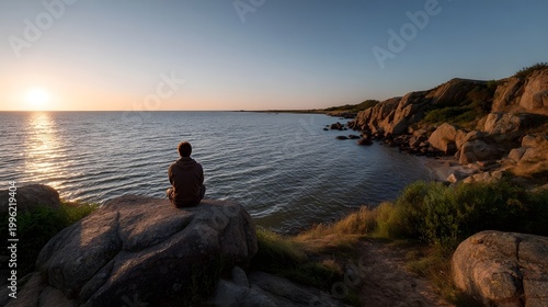 A solitary figure contemplates the vast ocean and golden horizon from a rocky coastal vantage point