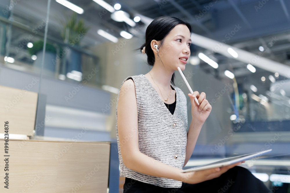 Fototapeta premium A young professional woman with a tablet and earphones sits thoughtfully in a modern office environment.