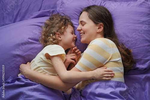 Canvastavla A mother and her child are lying close on a bed with purple sheets, sharing smiles and laughter