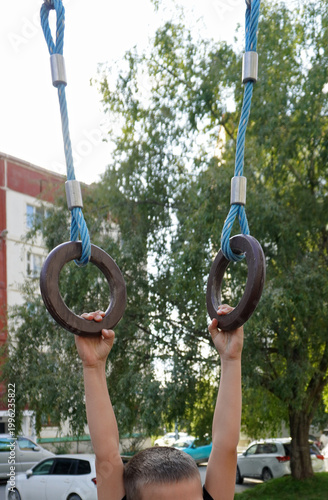 The child is hanging on gymnastic rings. A boy holding onto rings, seen from below, against the background of trees and parked cars in the courtyard.