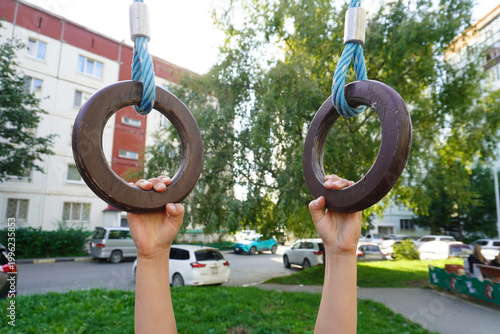 A child holds onto gymnastic rings in close-up. A child boy holds rings with his hands, against the background of a courtyard with houses and greenery.