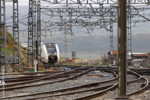 Commuters and travellers gather along the platforms at Ávila railway station as a regional train arrives, framed by overhead cables and urban architecture