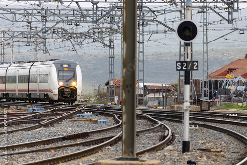 Commuters and travellers gather along the platforms at Ávila railway station as a regional train arrives, framed by overhead cables and urban architecture