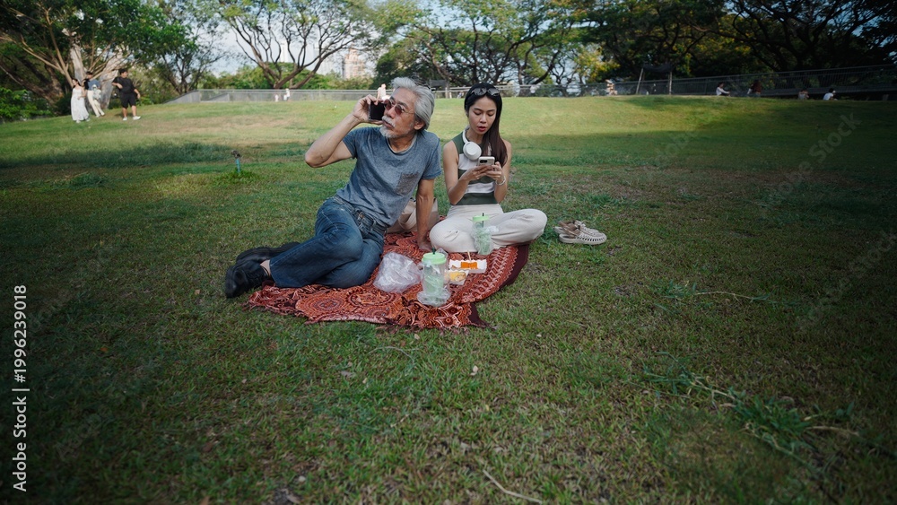 Fototapeta premium Senior man making phone call and young woman using social media during picnic in park