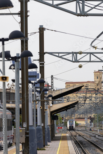 Commuters and travellers gather along the platforms at Ávila railway station as a regional train arrives, framed by overhead cables and urban architecture