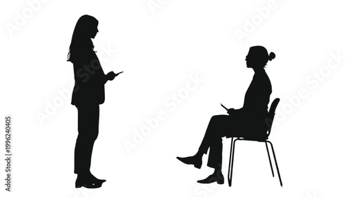 Businesswomen engage in a professional discussion while one stands and the other sits on a chair.