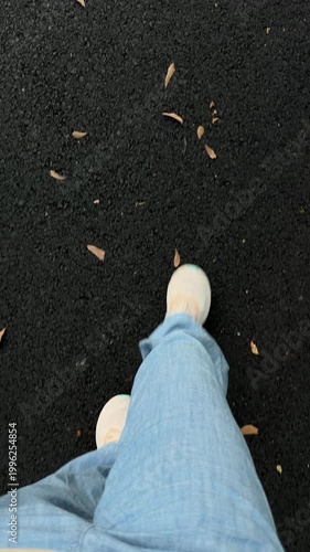 Handheld tracking shot of feet walking forward on asphalt path with scattered leaves in park during daytime autumn casual stroll