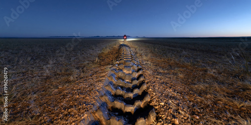 Low-angle shot of motorcycle tire tracks in Mongolian soil leading to a bright headlight under a June night sky