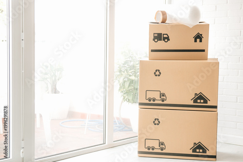 Schilderij op canvas Stack of cardboard moving boxes with truck and house icons, bubble wrap, and packing tape on top, against a white brick wall