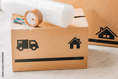 Canvastavla Stack of cardboard moving boxes with truck and house icons, bubble wrap, and packing tape on top, against a white brick wall