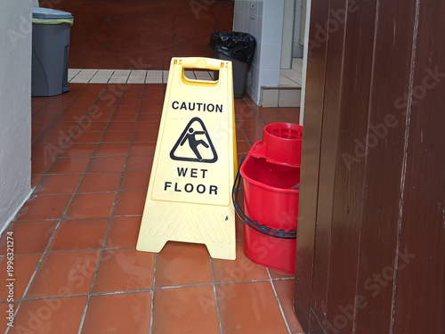 Caution wet floor sign in entrance to public conveniences, with a red mop bucket placed by side, ready to wash the toilet floors and keep the lavatories clean.