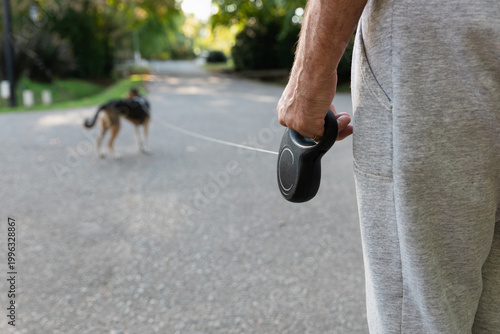 Close-up of a senior man's hands holding a retractable leash in a sunny neighborhood. Walking his dog on a bright afternoon with copy space. Elderly lifestyle and healthy activity
