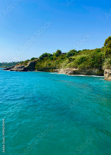 Beautiful turquoise sea with rocky coastline and green hills under clear blue sky. Summer seaside landscape with calm water and natural coastal scenery.