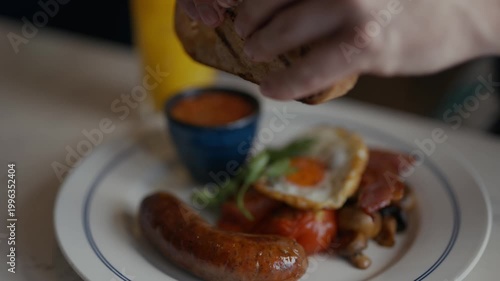 Cozy breakfast scene with sausages, eggs, and toast. Person's hands reach for food with gentle motion. Glass of orange juice adds warm, inviting color. Comforting meal suggests a relaxed morning start