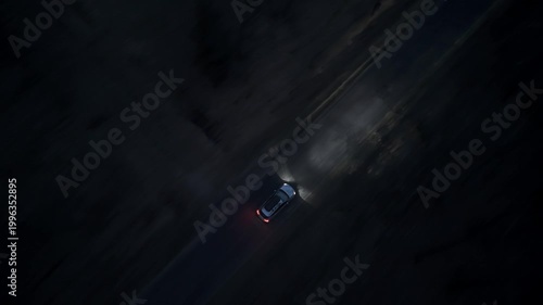 Drone follows a car moving through forest road at night with strong headlights illuminating narrow path in darkness