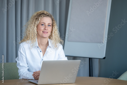 Business woman working at computer while sitting at office desk, blonde curly hair professional in neutral modern workspace, corporate environment with copy space.