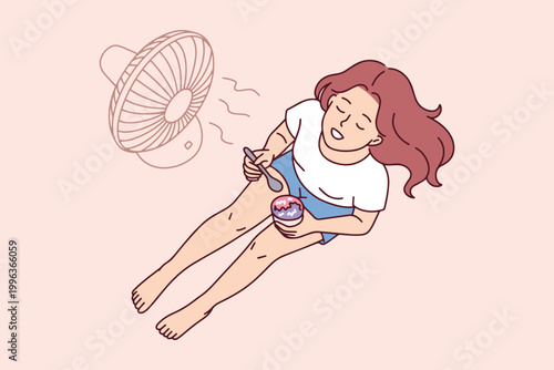 A young woman lies on the floor near an electric fan, eating cold fruit sorbet from a bowl to stay cool and comfortable during a summer heatwave at home.