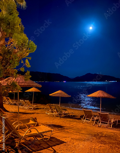 Night beach scene with sunbeds and umbrellas under bright moonlight. Calm sea with reflections and relaxing summer vacation atmosphere by the coast.