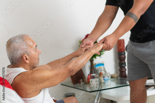 Young man helping elderly father stand up by holding hands showing physical support and care