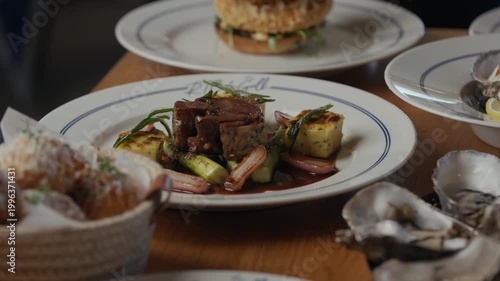 Wood table holds plates of food. Oysters rest beside a plated steak. Bread is nestled in a basket. Dinner looks rich and freshly prepared. Scene suggests a cozy, upscale dining experience