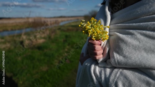 Close-Up of a Person's Hand Wrapped in a Soft Blanket Gently Holding a Small Bunch of Fresh Yellow Wildflowers Against a Serene Natural Landscape During Spring