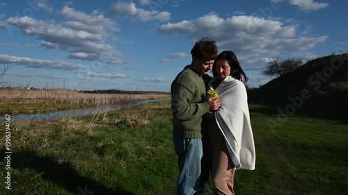 A Young Couple Shares a Tender Embrace and Romantic Moment by a Peaceful River in a Beautiful Green Field under a Blue Sky