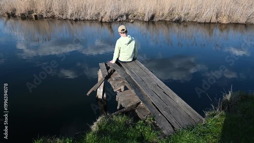 Young Man Sitting on a Dilapidated Wooden Pier by a Calm River, Reflecting and Enjoying the Serene Nature on a Sunny Day