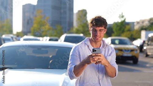 Smiling young man leaning on the car while holding mobile phone, typing message. Transport, technology and lifestyle concept. Real time