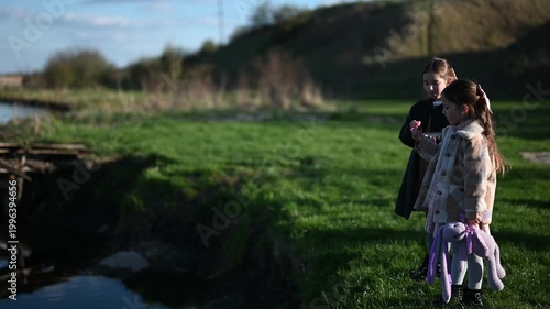 Two Young Girls Enjoy a Peaceful Outdoor Moment, Sharing Fresh Berries by a Grassy Riverbank with Their Beloved Stuffed Animal on a Sunny Spring Afternoon, Embracing Childhood Joy.