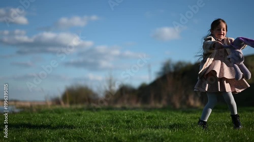 A Happy Little Girl with Long Hair Joyfully Spinning in a Green Field, Holding a Stuffed Bunny Toy Under a Bright Blue Sky on a Sunny Day