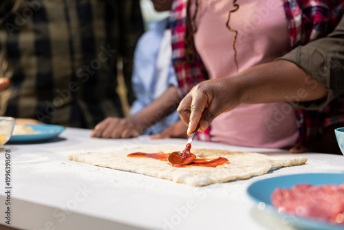 Close up of black kids using spoon to spread pizza sauce on the dough, making a homemade food recipe with fresh vegetables and toppings. Happy family cooking together at the kitchen island.