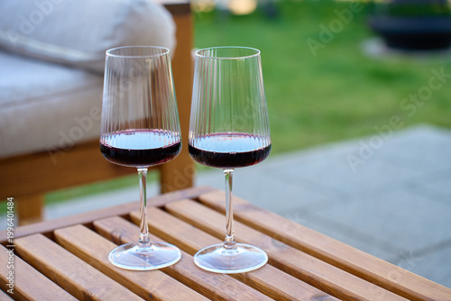 Two glasses with red wine stand on wooden table in outdoor patio setting