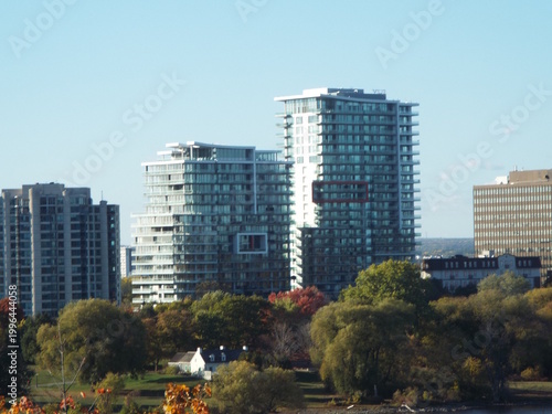 Modern glass high-rise apartment buildings tower over a lush, tree-filled landscape near a river in an urban setting.