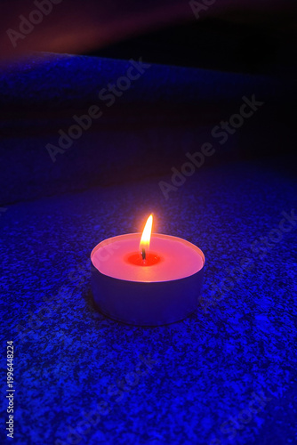 Burning tea light candle in blue night lighting. single candle flame glows orange against a dark blue grainy surface. The small flame