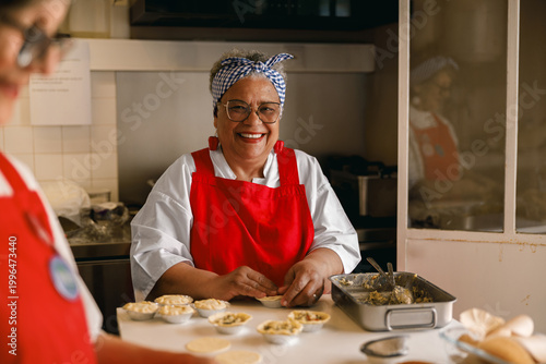 Foto A joyful senior woman of color looks directly at the camera while pressing dough into tart molds
