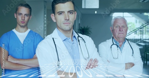 Standing center doctor crossing arms in clinic corridor with lab coats, blue scrubs, stethoscopes
