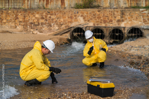 Εκτύπωση καμβά Environmental engineers in yellow hazmat suits collecting water samples from industrial drainage pipes for pollution and contamination testing