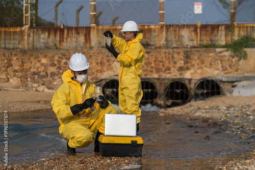 Canvas Print Environmental engineers in yellow hazmat suits collecting water samples from industrial drainage pipes for pollution and contamination testing