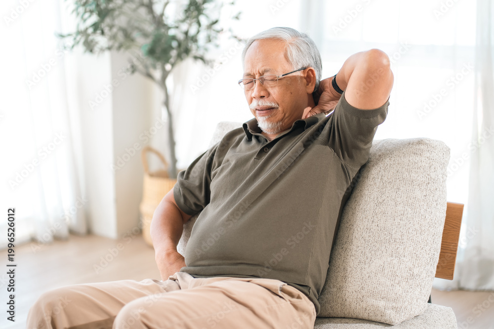 Obraz premium Elderly man with neck pain, sitting on sofa in bright room, discomfort and fatigue