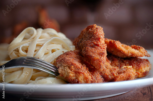 Milanesas de pollo pescado o camarones empanizados, en un plato blanco con tallarines o pasta fetuccini, con tenedor al centro y con carne de res al fondo.