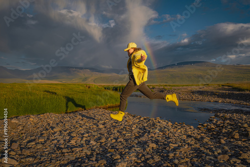Obraz na plátně Young woman in a yellow jacket and boots jumping over a stream in a mountain valley under a dramatic sky with a rainbow