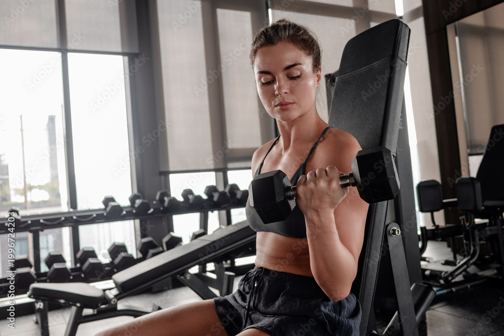 Fototapeta premium Athletic woman performing seated dumbbell bicep curl on incline bench in modern gym