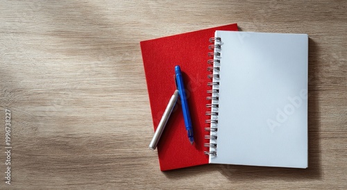 A red notebook and a white spiral-bound notepad rest on a light wood surface with two pens.