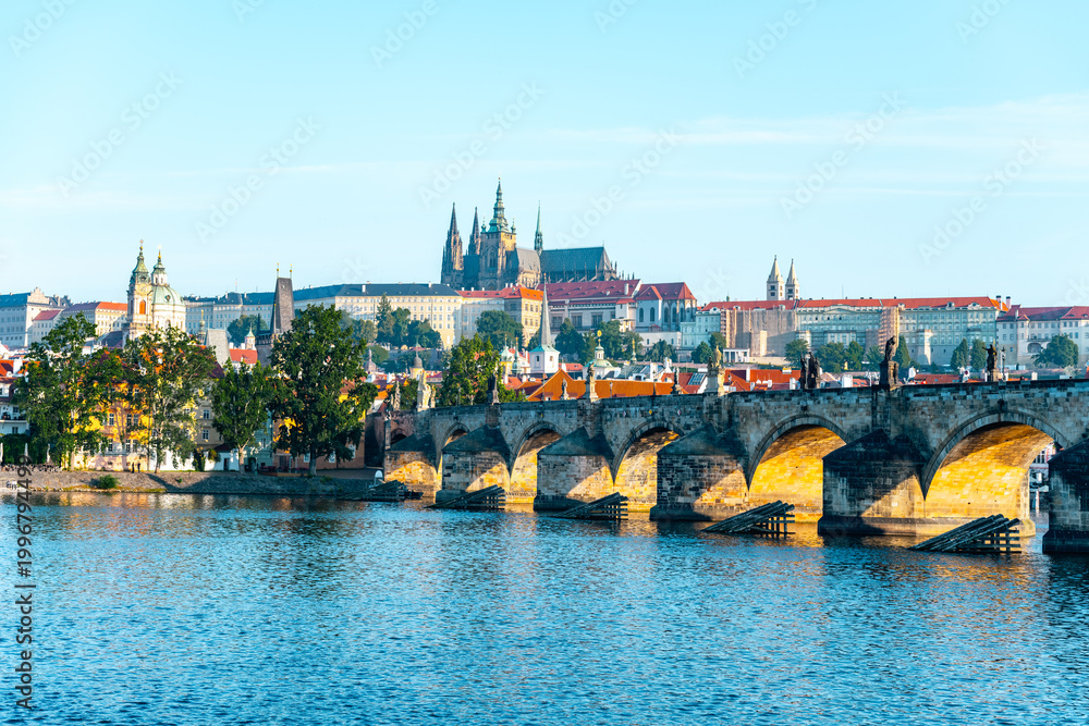 Obraz premium Stunning panorama of Prague with the historic Charles Bridge arching over the Vltava River, framed by the majestic Prague Castle under a clear blue sky, showcasing the city's rich architecture.